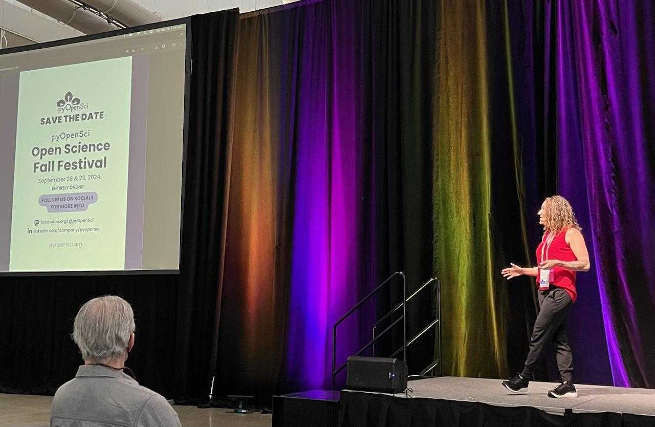 Image of a woman on a big stage wearing a red tank top talking. On the side is a slide that says pyOpenSci - Save the Date. Open science fall festival september 28-29 2024