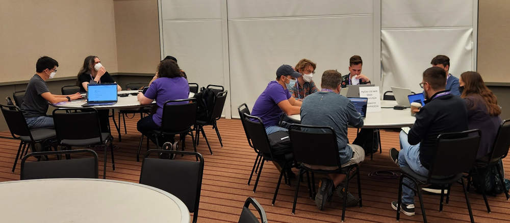 Image showing a room with round tables and people working at each table most of who have laptops sitting in front of them.