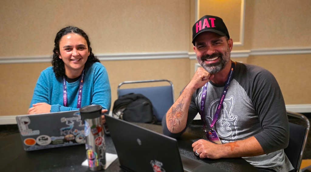 Two smiling PyCon US 2026 attendees sitting at a round table with laptops during a conference sprint. Both wear purple PyCon US 2026 lanyards from Long Beach.