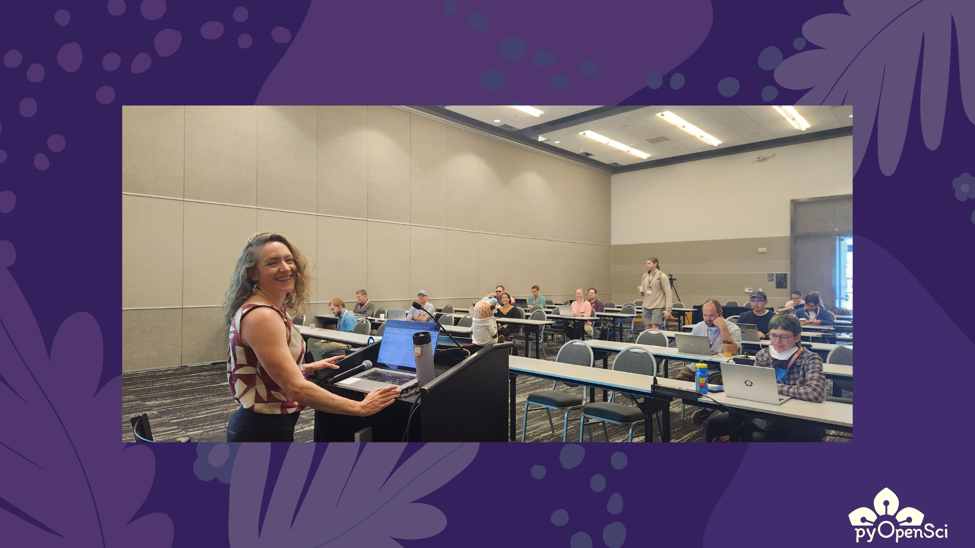 A woman Leah Wasser, smiling at the podium while preparing to present at a pyOpenSci workshop at SciPy 2025, with attendees gathered behind her.