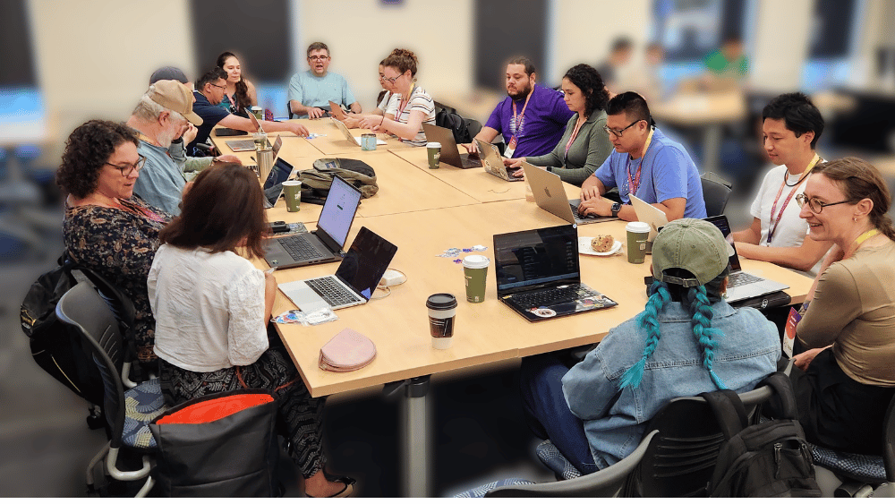 A group of contributors sitting at a table at a pyOpenSci sprint working on their laptops and talking to each other.